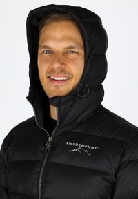 Svart puffjacka med huva, med dragkedjestängning och logotypen "SWEDEMOUNT". Tyget är slätt med ett kviltat mönster.