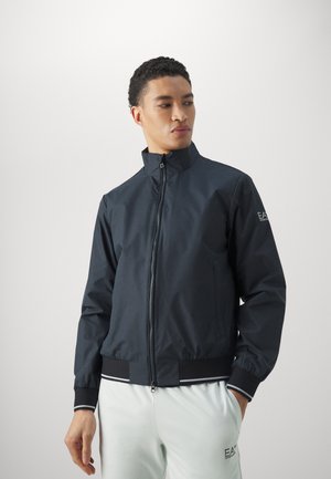Giacca bomber blu navy con colletto alto, chiusura con zip e polsini elasticizzati. Presenta un logo sulla manica e dettagli a righe all'orlo.