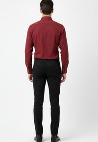 Hombre de pie de espaldas, vestido con una camisa de manga larga burdeos, pantalones ajustados negros y zapatos negros, frente a un fondo blanco liso.