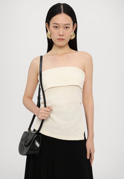 Cremfarbener trägerloser Top aus strukturiertem Stoff mit überlappendem Design, kombiniert mit einer schwarzen glänzenden Tasche mit geschwungenem Klappdeckel und Schultergurt.