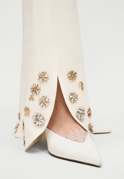 Κοντινή λήψη σε παντελόνια σε κρεμ απόχρωση με χρυσές και ασημί ανθικές διακοσμήσεις, συνδυασμένα με λευκές μυτερές γόβες και κάλτσες από δαντέλα.