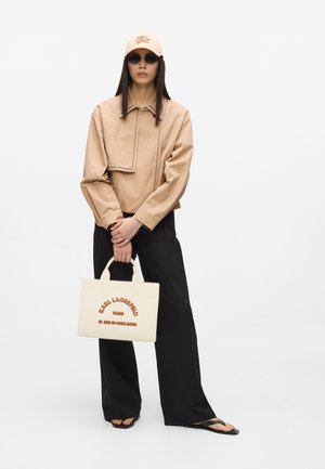 Ženska, oblečena v bež kratko jakno, ujemajočo kapo, črne sončna očala, široke črne hlače, japonke, drži belo torbo Karl Lagerfeld.