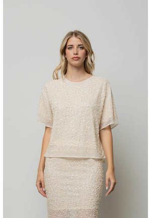 Donna con lunghi capelli biondi che indossa una maglietta beige a maniche corte strutturata e una gonna beige fino al ginocchio su uno sfondo grigio uniforme.