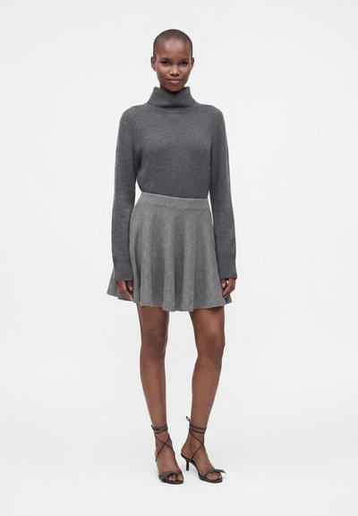 Femme debout portant un pull à col roulé gris foncé, une jupe plissée grise et des sandales à talons à brides noires sur un fond blanc uni.