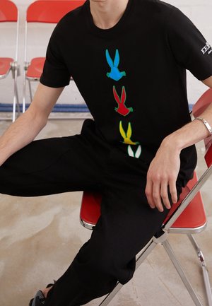 T-shirt noir avec quatre silhouettes de lapins stylisées et multicolores (bleu, vert, rouge, jaune) disposées verticalement. Matière en coton.
