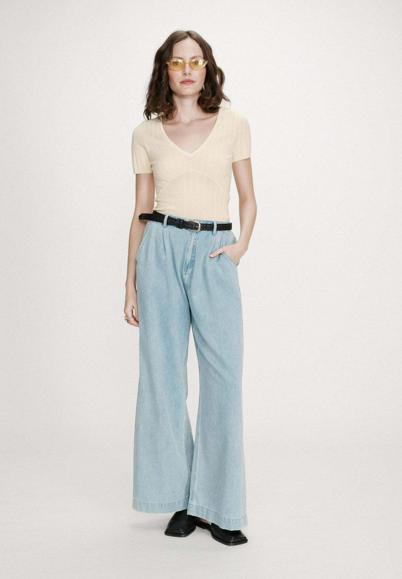 Femme debout portant un haut beige à manches courtes et encolure en V, un jean large bleu clair avec ceinture noire, des chaussures noires et des lunettes à verres jaunes.