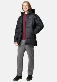 Giubbotto piumino nero con cappuccio, caratterizzato da un design trapuntato, zip frontale e tasche laterali. Indossato sopra una maglietta bordeaux e pantaloni grigi.