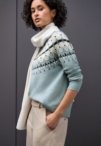 Maglione lavorato a maglia azzurro chiaro con motivi geometrici e accenti scuri; abbinato a una sciarpa crema e pantaloni beige con texture.
