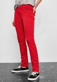 Jeans skinny de mezclilla roja con una textura suave, cinco bolsillos y cierre de botón. Combinados con zapatillas negras y blancas.