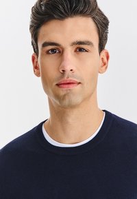 Granatowy sweter dziany z okrągłym dekoltem, z białym wewnętrznym kołnierzem. Gładka faktura i dopasowany krój. Modelka z włosami o średniej długości.