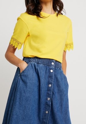 Żółta bawełniana koszulka z koronkową lamówką na rękawach, zestawiona z niebieską spódnicą jeansową z elastycznym paskiem w talii i ozdobnym guzikiem z przodu.