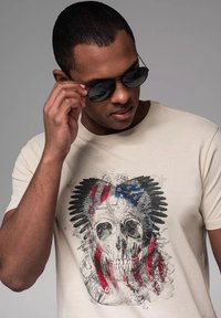Beige bomulds t-shirt med grafisk design af et kranium med røde og blå accenter. Model iført sorte solbriller, teksturerede stofdetaljer synlige.
