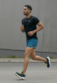 Runner con una maglietta nera a maniche corte e pantaloni corti color turchese, che indossa scarpe da corsa blu navy con dettagli verdi. Sfondo di asfalto, bassa vegetazione.