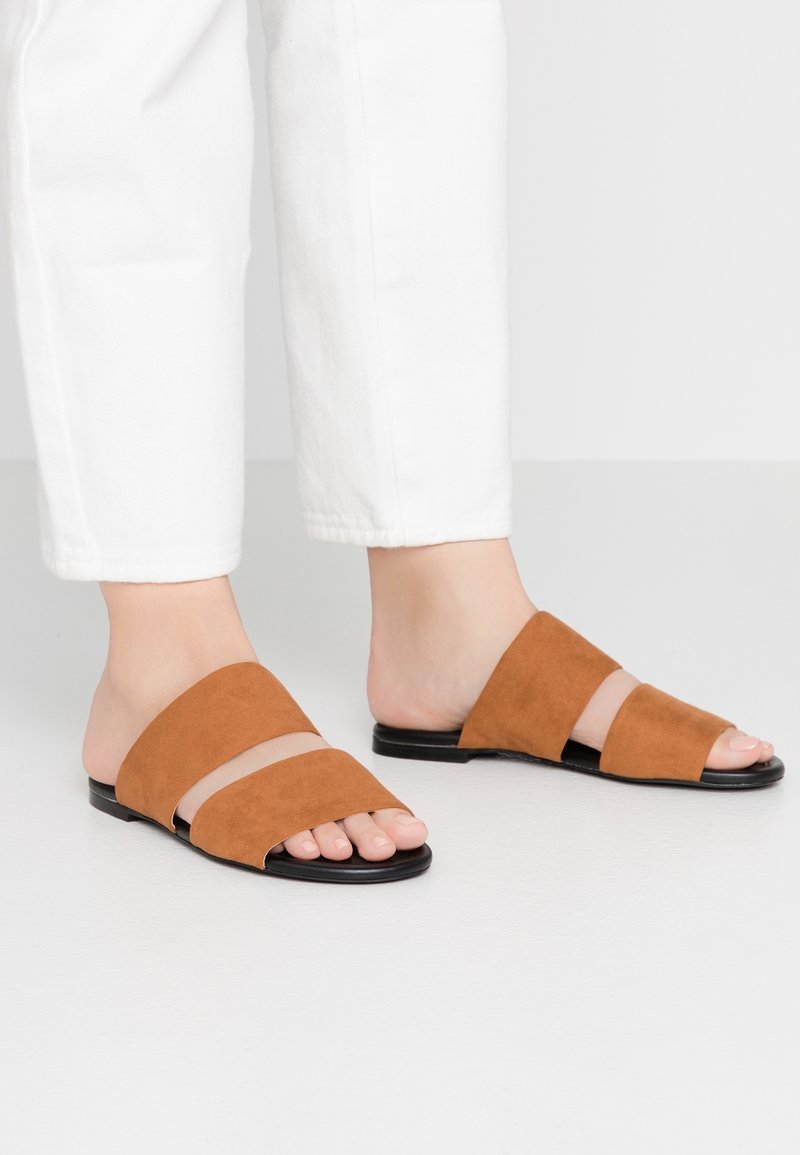 Pieds chaussés de sandales marron en daim à enfiler avec deux larges brides et semelles noires, portées avec un pantalon blanc coupé au dessus de la cheville.