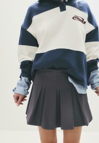 Person, die einen navyblauen und weißen Sweatshirt mit dem Text "Stockholm" trägt, ein blau gestreiftes Hemd darunter und einen gefältelten dunkelgrauen Rock.