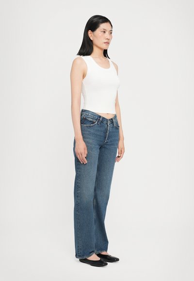 Débardeur blanc à côtes, jean en denim bleu taille haute et ballerines noires. La mannequin se tient de profil, mettant en valeur l'ajustement et le design de la tenue.