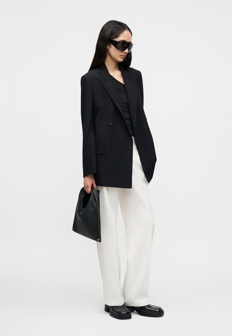 Blazer noir croisé sur un haut noir, pantalon blanc à jambes larges, sac noir et grandes lunettes de soleil noires. Textures lisses et design ajusté.