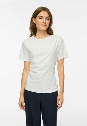 Jeune femme aux cheveux châtain clair porte un haut blanc uni à manches courtes et un pantalon bleu foncé, debout devant un fond blanc.