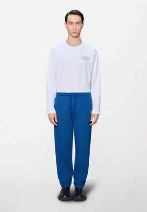 Ung mann som står rett opp og ned, iført en hvit langermet skjorte med en liten logo og blå bukser med snøring, samt svarte sko.