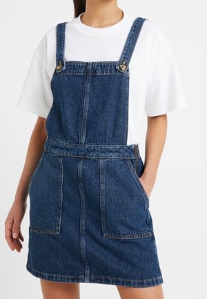 Kvinde iført en blå denim-forklædekjole over en enkel hvid t-shirt med korte ærmer, med den ene hånd i kjolens lomme.