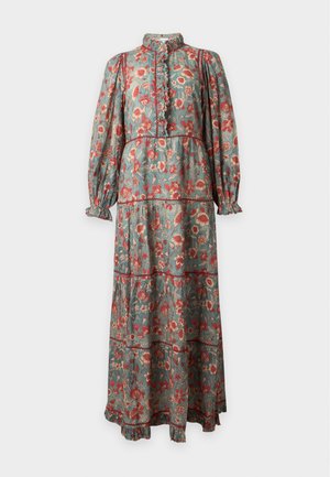 Femme debout portant une longue robe verte à motif floral avec des manches longues et des chaussures plates beiges sur un fond clair uni.