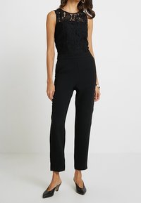 Mono negro con parte superior de encaje sin mangas con patrones florales, un cuerpo texturizado y pantalones ajustados. Combinado con tacones negros.
