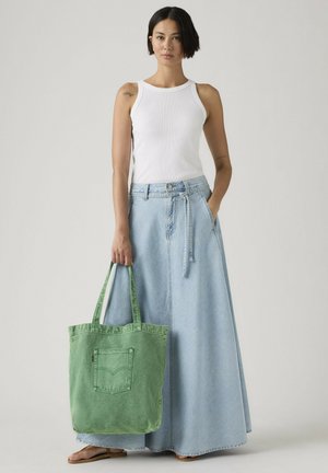 Kobieta w białym topie bez rękawów i długiej, jasnoniebieskiej dżinsowej spódnicy, trzymająca zieloną torbę na ramię i mająca na stopach brązowe klapki, stojąca na jednolitym tle.