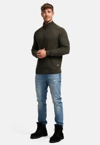 Sudadera de forro polar verde oliva con cuello de cremallera y puños acanalados, combinada con jeans desgastados de color azul claro y botas negras con cordones.