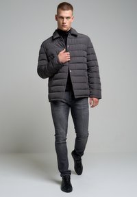 Šedá puffer bunda s horizontálním prošíváním, zapínání na knoflíky a límec, nošená přes černý rolák a spárovaná s tmavými džíny a černými botami.