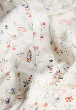 Weißer Stoff mit intrikaten Mustern, die bunte Blumen, Vögel und Formen zeigen. Weiche Textur mit sichtbaren Nähten und Knopfdetails.