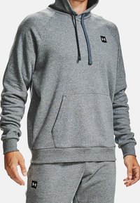 Felpa grigia con cappuccio, dotata di tasca frontale, polsini e orlo a coste, caratterizzata da un cordino blu e un piccolo patch con logo nero.