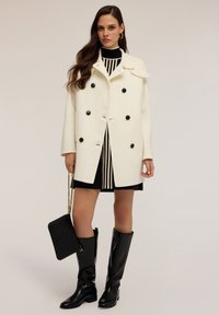Cappotto bianco doppiopetto con bottoni neri, indossato sopra un vestito a righe. Stivali neri al ginocchio e una borsa nera strutturata. Finitura in tessuto liscia.