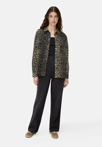 Frau mit langen braunen Haaren, die eine Jacke mit Leopardenmuster, ein schwarzes Oberteil, weite schwarze Jeans und beigefarbene Sneakers trägt und vor einem schlichten Hintergrund steht.
