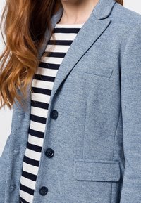 Vaaleansininen bleiseri teksturoitua kangasta, yksirivinen malli kahdella napilla, lovilla varustetut kaulukset ja rintatasku. Raidallinen paita alla.