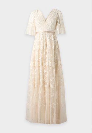 Femme aux longs cheveux noirs portant une robe beige en dentelle longue jusqu'au sol avec broderies florales et manches mi-longues, debout devant un fond uni.