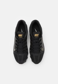 Sapatilhas de treino preta da Nike com um design texturizado, apresentando um logo dourado em destaque e detalhes reflexivos nos lados e nos atacadores.