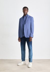 Light blue blazer over a dark blue shirt, paired with gradient blue jeans and white sneakers. Smooth fabric with tailored design.