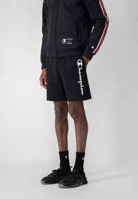 Giacca sportiva nera con strisce rosse e bianche, abbinata a pantaloni corti neri con il logo "Champion" bianco. Sneakers nere e calzini.