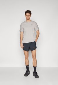 Light gray short-sleeve t-shirt with blue logo, paired with dark shorts featuring orange Nike logo. Black athletic shoes and socks complete the look.