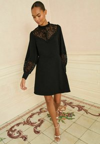 Črna obleka z čipkastim korpusom in rokavi, s prilegajočo silhueto in nagubanimi ovratniki. Tkanina je gladka s zapletenimi čipkastimi detajli.