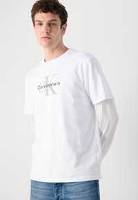 Joven con cabello rizado que lleva una camiseta blanca con el logo de Calvin Klein sobre una camisa blanca de manga larga y vaqueros azules, con un fondo liso.