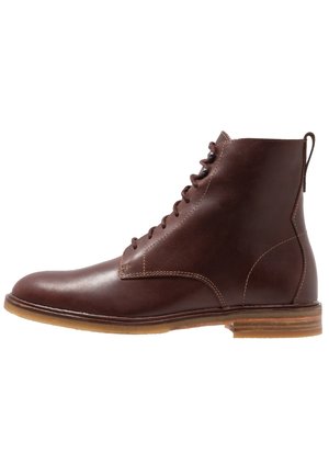 Brauner Leder-Stiefelette mit Schnürsenkeln vorne, runder Spitze und flacher Holzsohle mit leichtem Absatz, vor weißem Hintergrund.