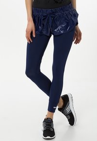 Marina leggings med hög midja, i kombination med en blank, marina, shorts med dragsko. Modellen bär svarta träningsskor.