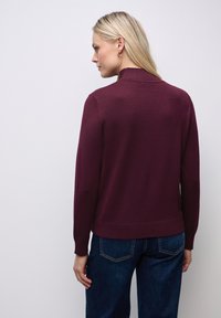 Bordeaux strikket sweater med høj krave, ribbede ærmer og liv, parret med mørkeblå jeans; glat tekstur og tætsiddende design.