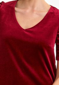 Top in velluto bordeaux con scollatura a V e maniche a puff corte. Texture liscia e design aderente, con una leggera lucentezza e senza chiusure visibili.