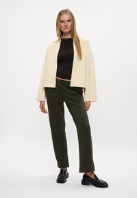 Kremowa kurtka z warkoczowym splotem, czarna dopasowana bluzka, oliwkowe dresy i czarne loafersy. Proste faktury w luźnym kroju.