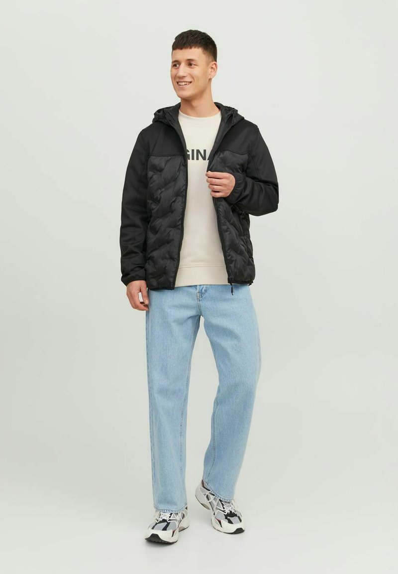 Zwarte gewatteerde jas met een capuchon, contrasterende gladde mouwen, gedragen over een licht sweater en gecombineerd met lichtblauwe jeans en sneakers.