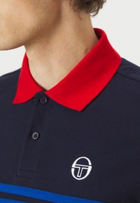 Tengerész kék póló piros gallérral, kék csíkokkal a mellen és fehér logóval. Az anyag textúrája látható, gombos részletekkel.