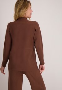 Bruine, lange mouwen, geribbelde gebreide top met een hoge hals en zijsplitten, gecombineerd met bijpassende bruine broek. Glad textuur en een comfortabele pasvorm.