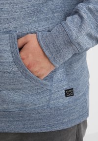 Blue blended fabric sweatshirt with a front pouch pocket, ribbed cuffs and hem, and a small black label reading “BLEND.”